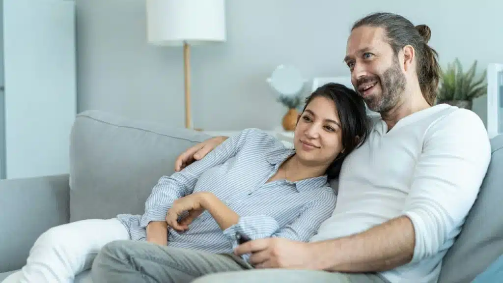A man and a woman are sitting close together on a sofa, looking right and smiling.