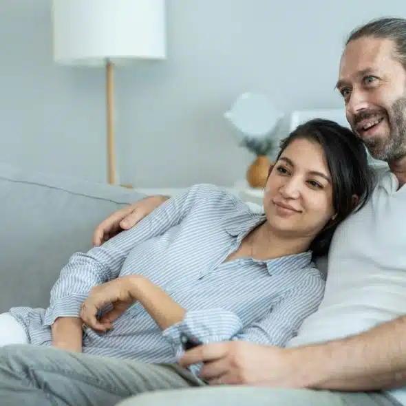 A man and a woman are sitting close together on a sofa, looking right and smiling.