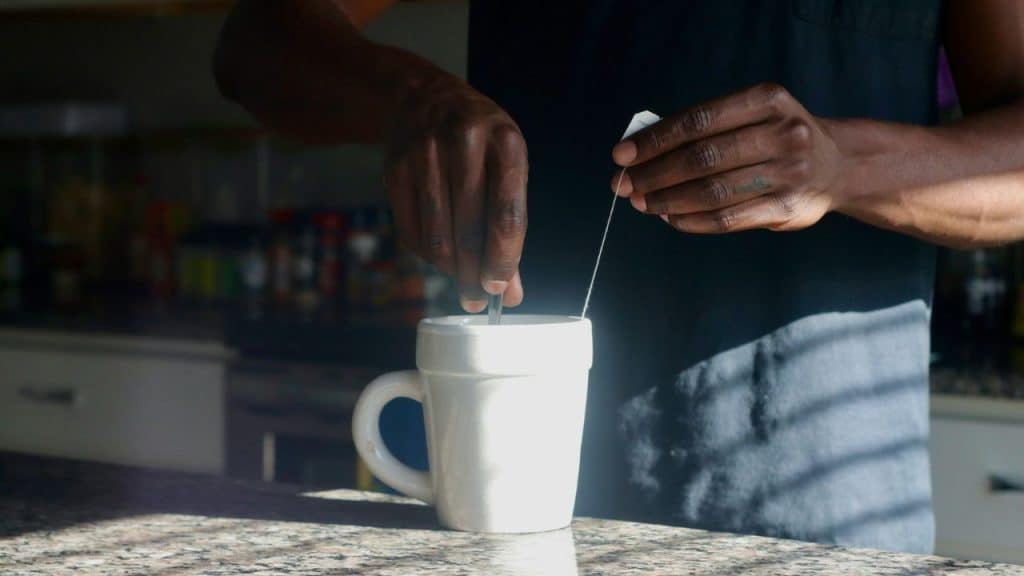 A close-up of a man's hands putting a tea bag into a white mug on a counter.