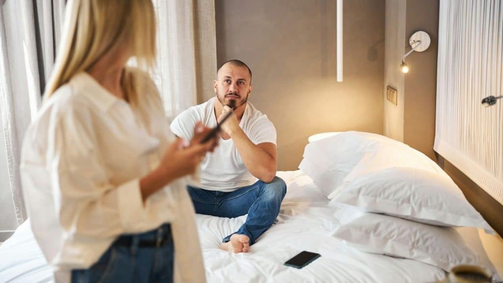 A man sits on a bed, looking seriously at a woman standing and talking.