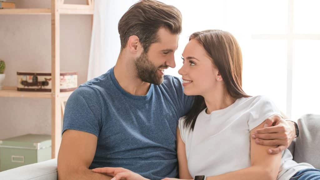 A man and woman cuddling on a light gray couch, smiling and looking off-camera.