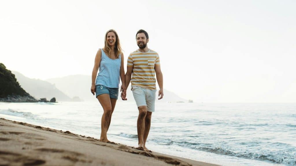 A smiling couple walks barefoot, holding hands on a sandy beach by the ocean.