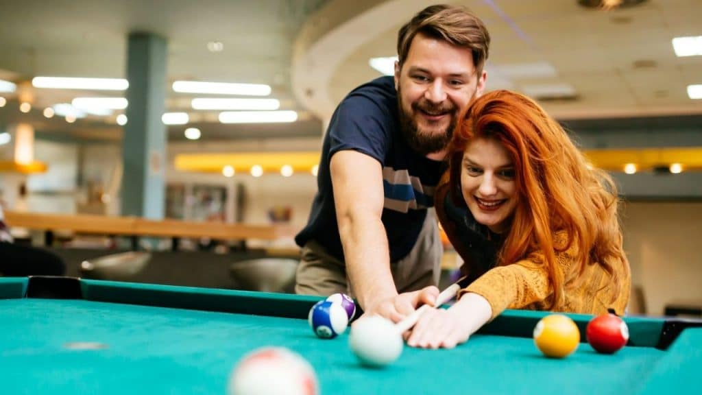 A smiling man leans over a woman with red hair as she lines up a pool shot.