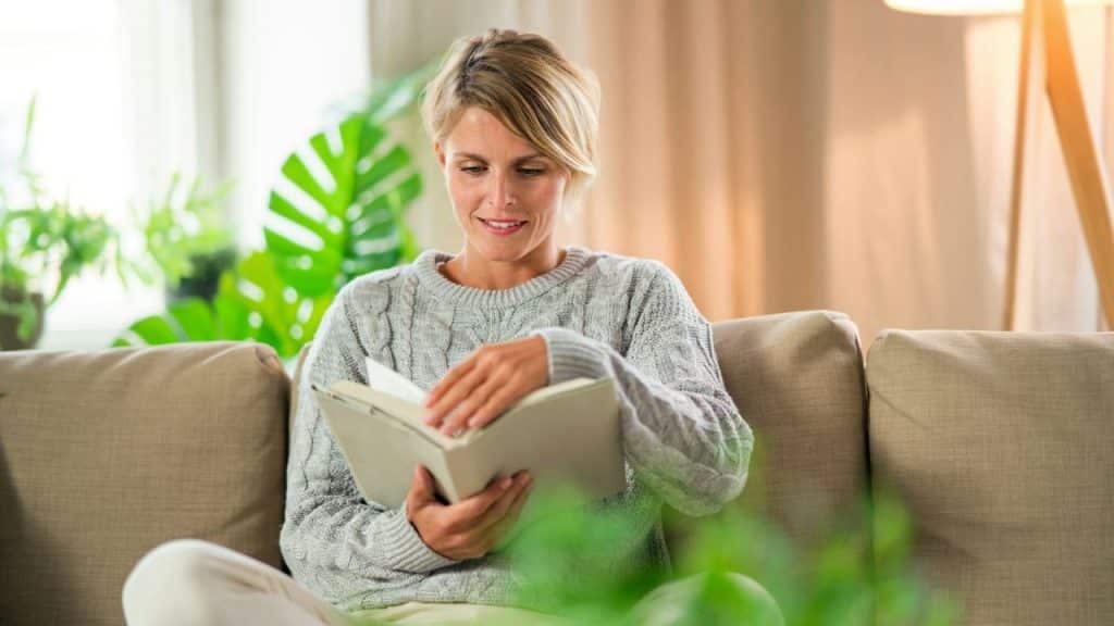 A woman in a gray sweater sits on a couch, smiling and reading a book.