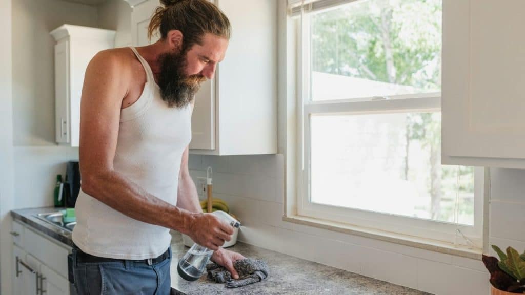 A man in a tank top cleans a kitchen counter next to a bright window.