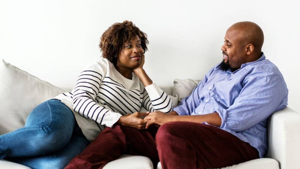 A smiling couple sits on a couch, holding hands and engaging in conversation.