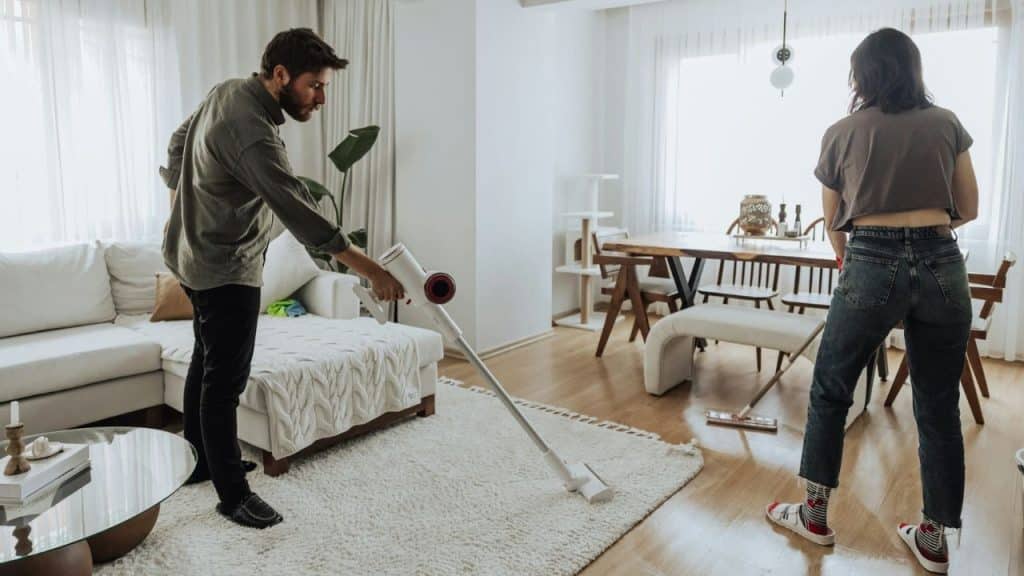 A man uses a stick vacuum to clean a white rug while a woman cleans the wooden floor behind him.