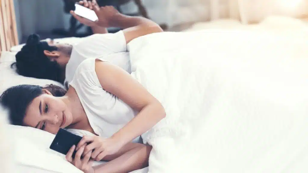 A man and woman lie in bed under white covers, both looking at their phones.