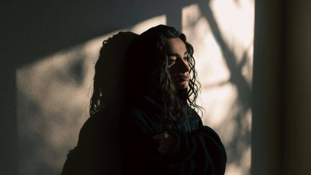 A woman with dark, curly hair stands in shadow, looking away with a melancholic expression.