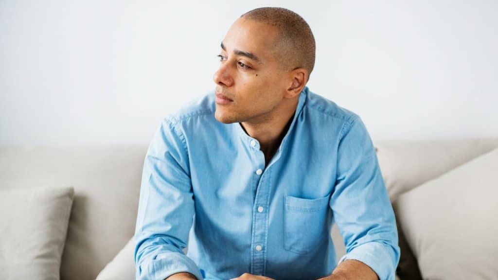 A man in a blue shirt with a shaved head sits on a couch, looking away thoughtfully.