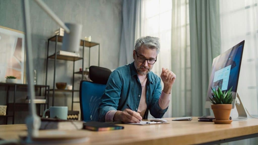 A middle-aged man with glasses writes in a notebook at a modern office desk.