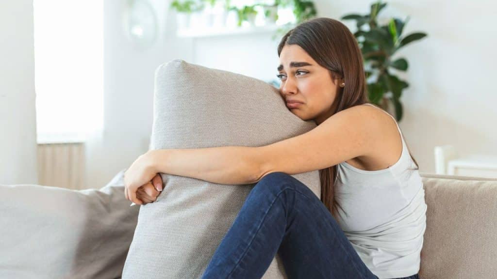A woman on a couch hugs a pillow tightly, looking distressed with tears in her eyes.