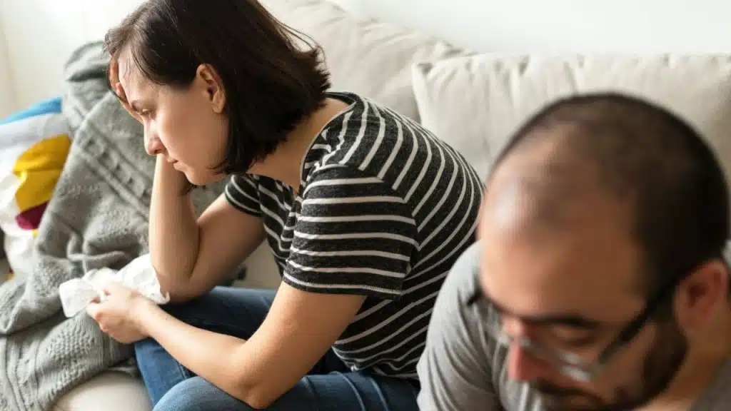 A woman in a striped shirt sits on a couch, holding her head, with a man out of focus.