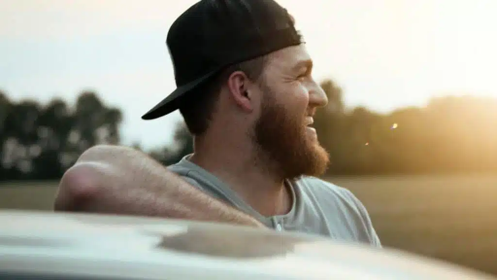 A bearded man in a backward cap smiling outdoors in warm evening light.