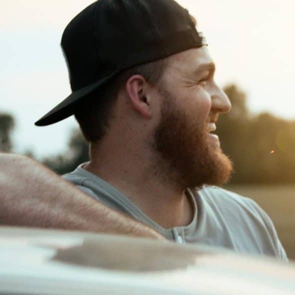 A bearded man in a backward cap smiling outdoors in warm evening light.