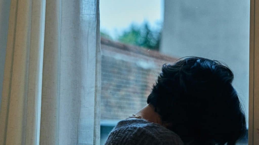 A person with short dark hair sits by a window, looking outside through sheer curtains.