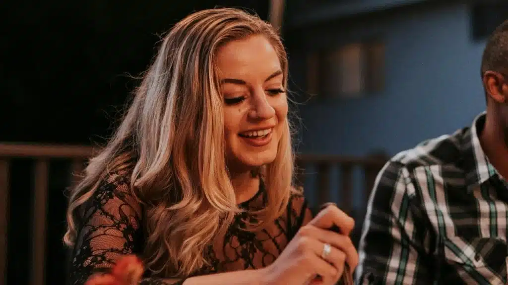 A smiling woman with long blonde hair sitting at a dinner table outdoors.