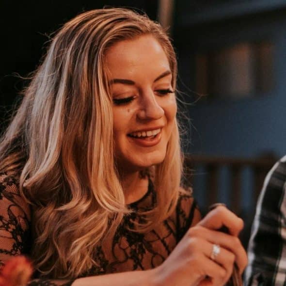 A smiling woman with long blonde hair sitting at a dinner table outdoors.