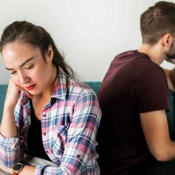 A woman and man sit back-to-back on a teal couch, looking upset or distant.