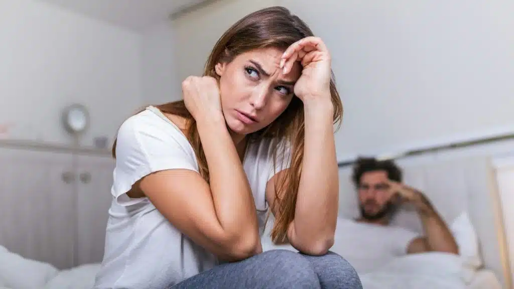 A distressed woman sits on a bed, holding her head, with a blurry man behind her.