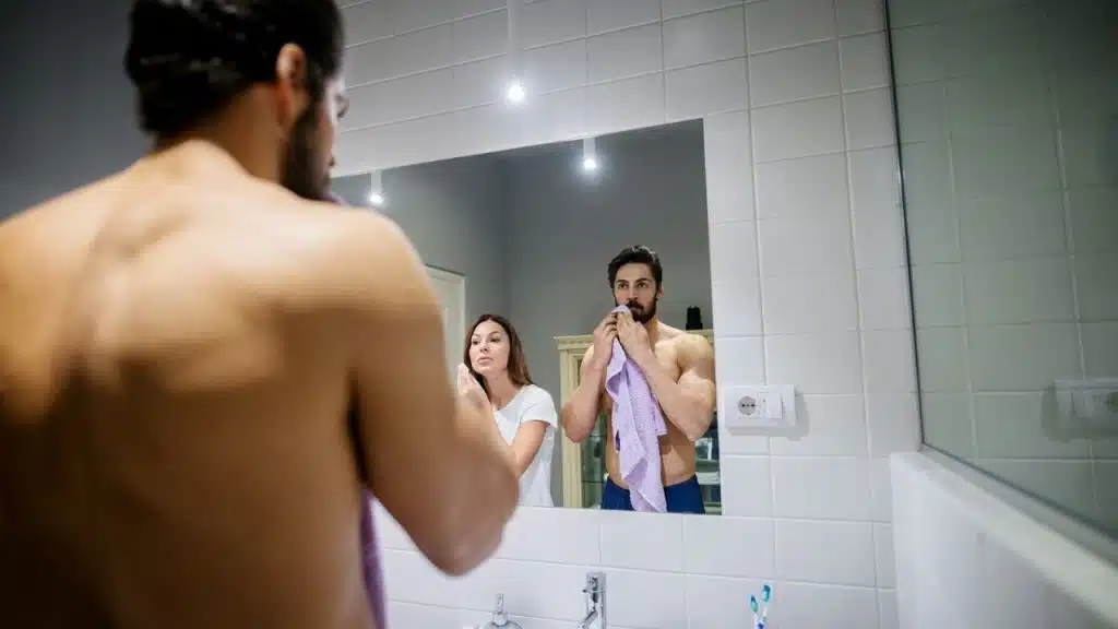 A man looks in a bathroom mirror while a woman stands behind him, also looking.