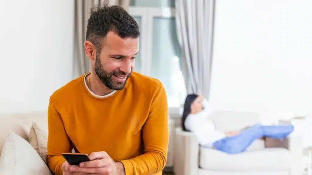 A smiling man uses a phone while a woman relaxes on a sofa in the background.