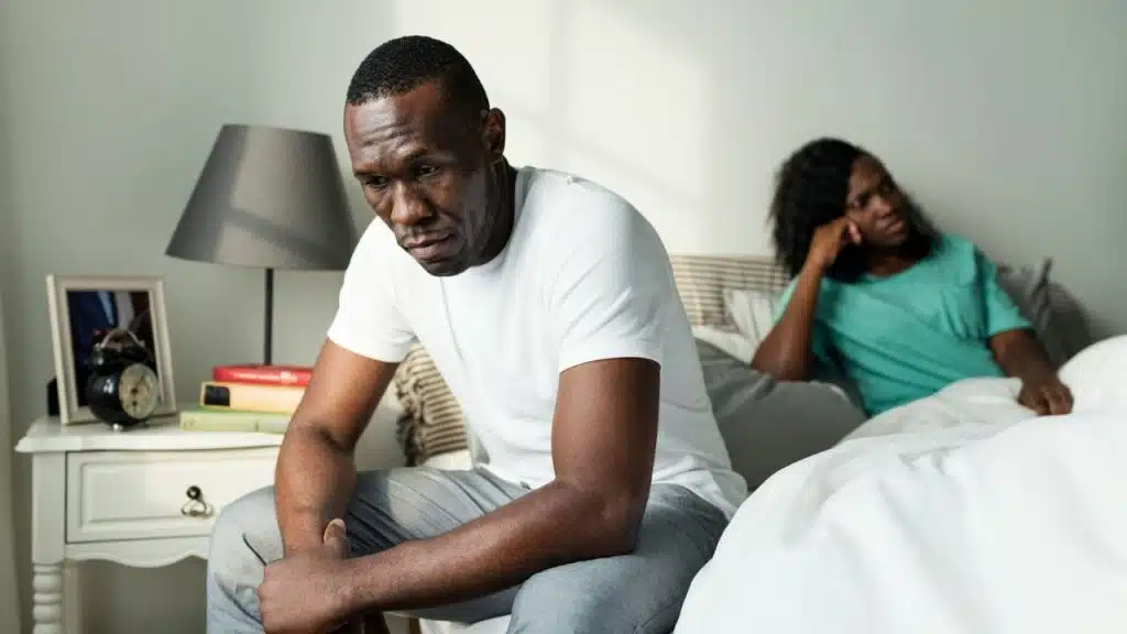 A man sits looking sad on a bed with a woman sitting frustrated in the background.