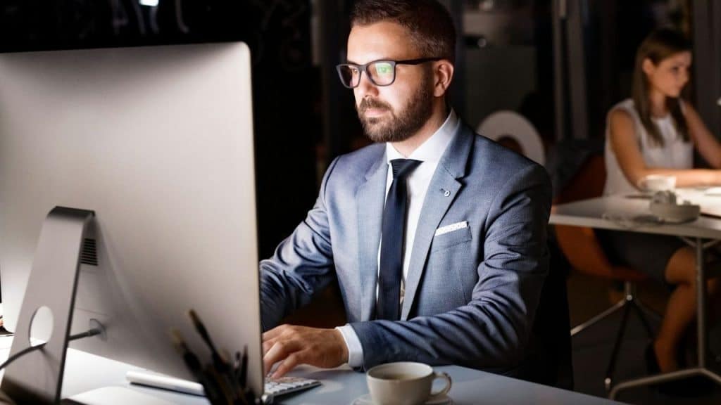 A man in a suit and glasses works on a computer late at night in an office.