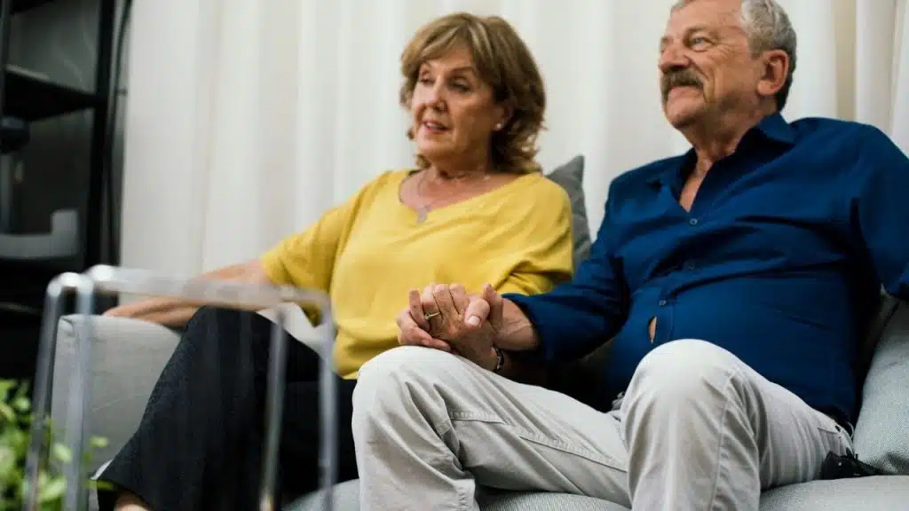 Elderly couple sitting on a couch, holding hands, and looking off to the side.