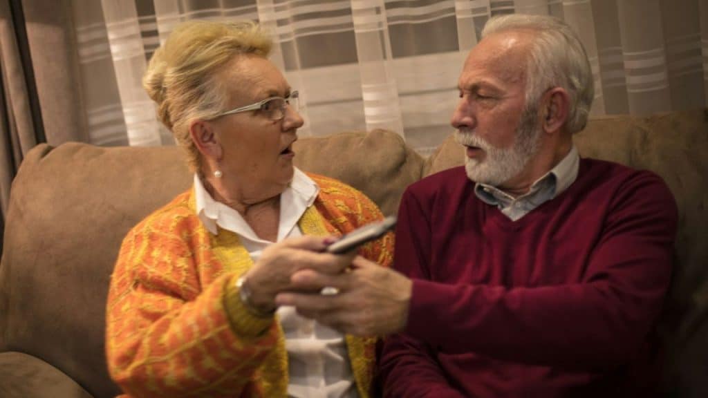 Elderly couple on couch, woman in yellow/orange cardigan holding remote, man in red sweater.