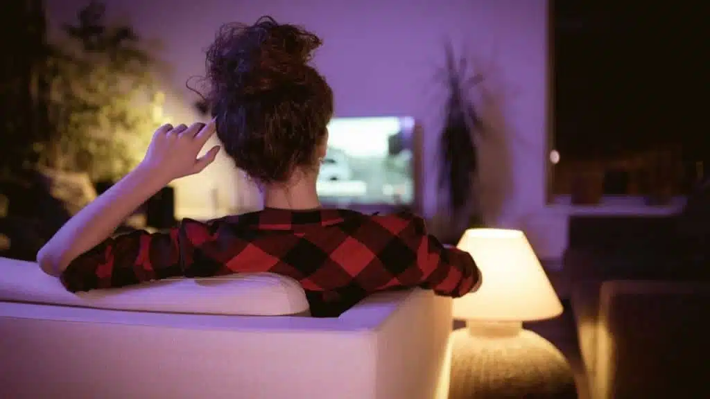 Woman in plaid shirt sits on couch watching TV in dimly lit, purple room.