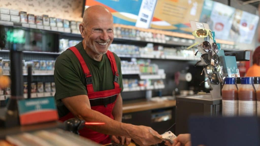 Smiling gas station attendant in red overalls and green shirt takes money from a customer.