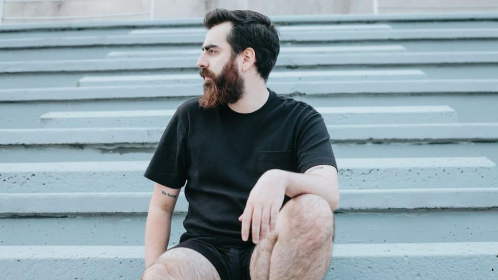 Young man with beard and tattoos in black shirt sits on grey stadium steps.