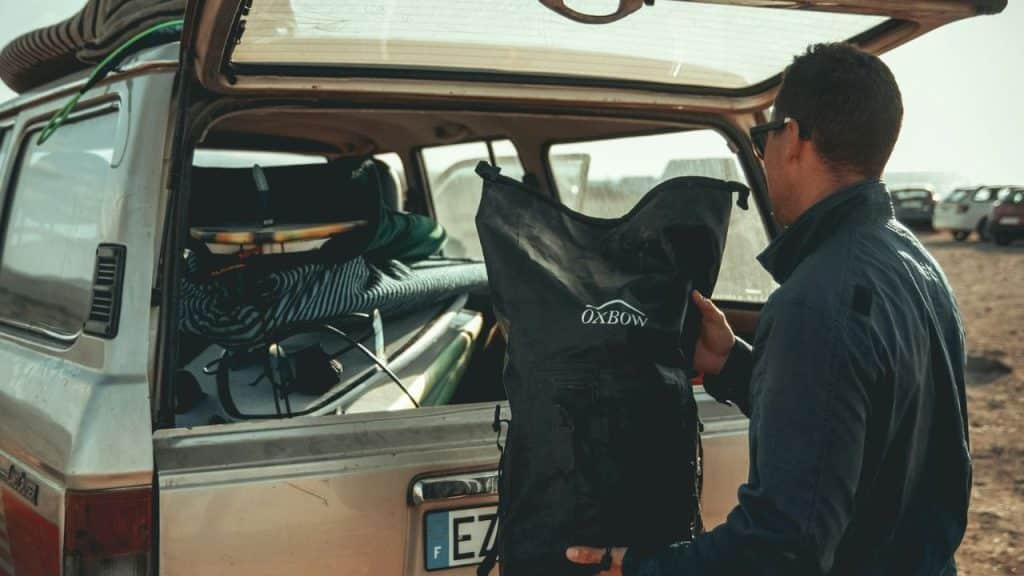 Man loads black "OXBOW" dry bag into crowded car trunk with surfboards.