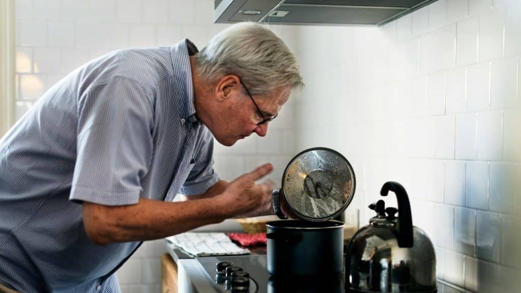 Elderly man in light shirt leans over stove to look into cooking pot.