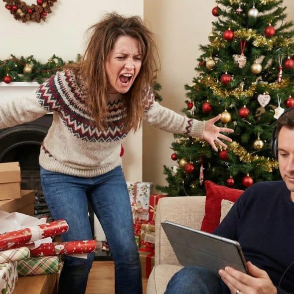 Stressed woman screams at man with headphones and tablet in Christmas room.