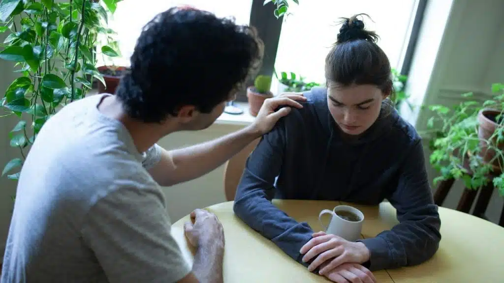 A man comforting a sad-looking woman by placing his hand on her shoulder at a table indoors.