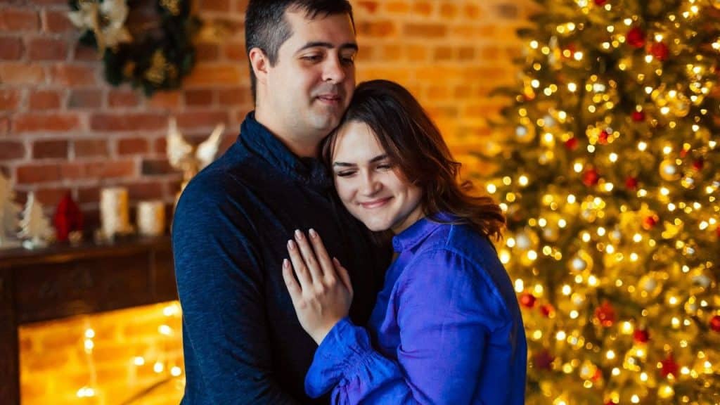 A smiling couple embracing near a brightly lit Christmas tree and a brick fireplace.