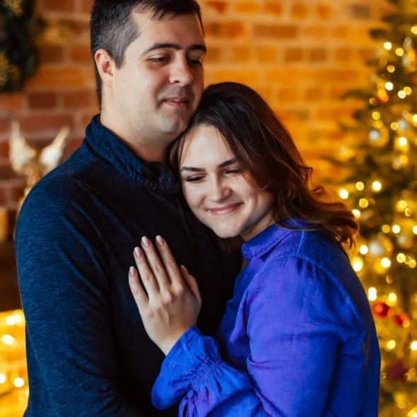 A smiling couple embracing near a brightly lit Christmas tree and a brick fireplace.
