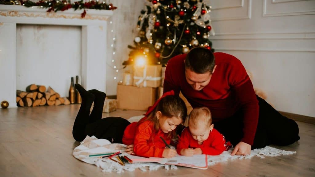 A father and his two young children are coloring on the floor by a Christmas tree.