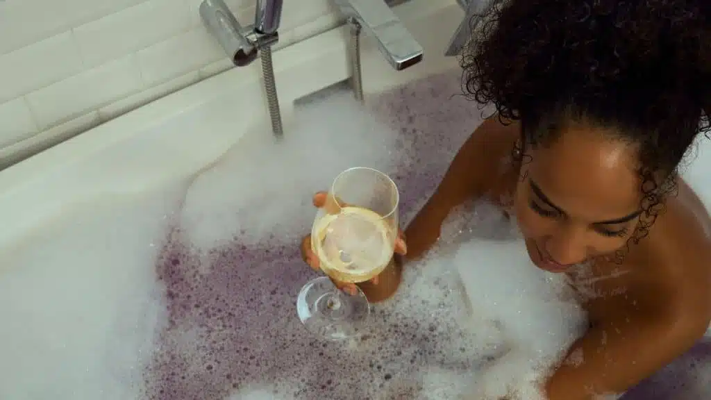 A smiling Black woman in a bubble bath holding a glass of white wine.