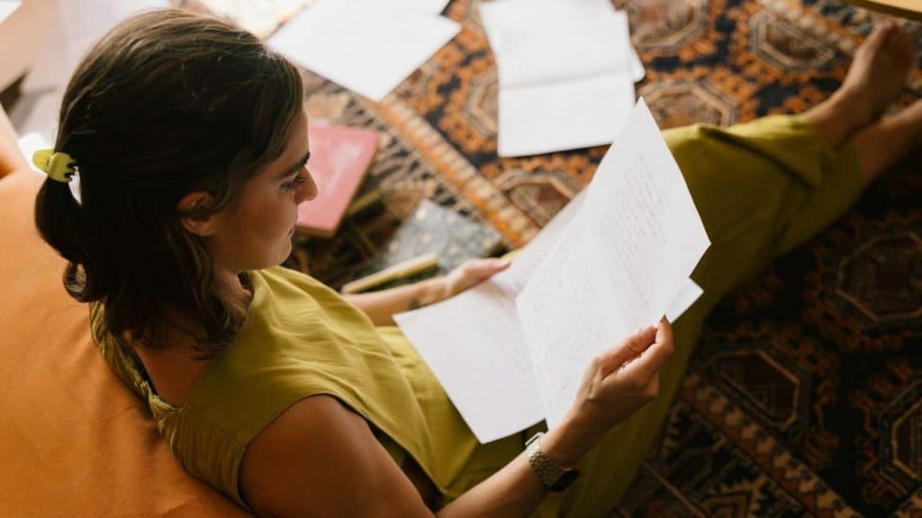 A woman sitting on a rug reads a paper while relaxing indoors with her feet up.