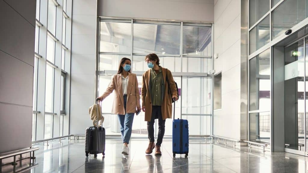 A couple wearing face masks walks through an airport with rolling suitcases.