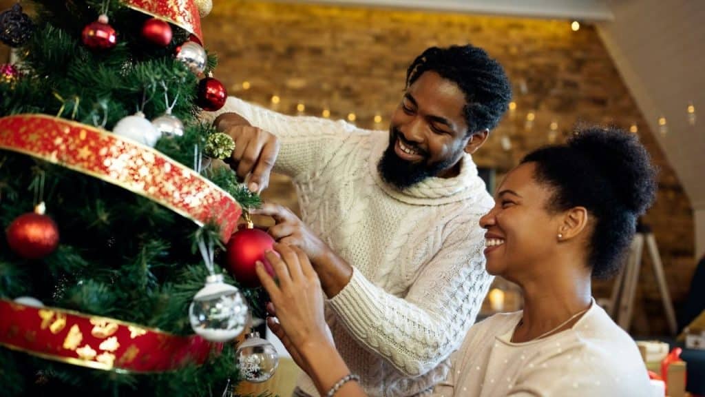 A smiling Black couple decorating a Christmas tree with red and white ornaments