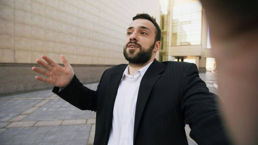 Man in suit and white shirt gestures and speaks outdoors in a public square.
