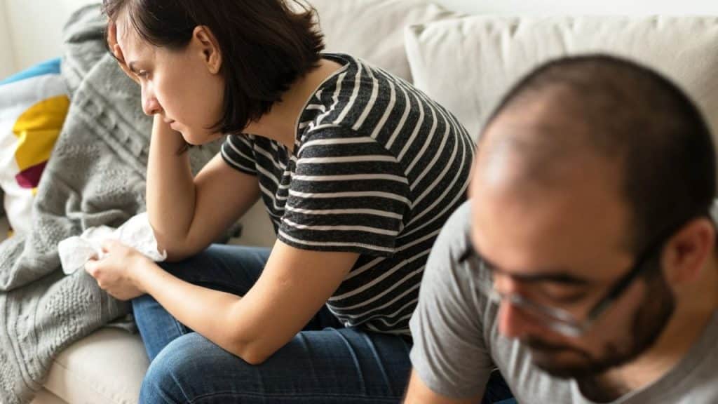 A distressed woman sits holding a tissue with a blurred man in the foreground.