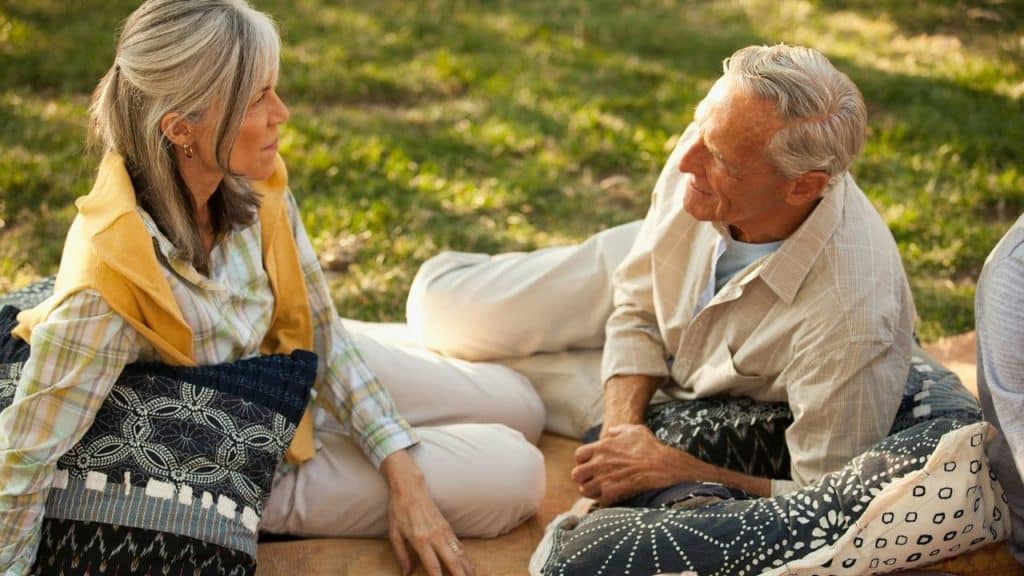 Elderly couple sitting on patterned pillows on a blanket outdoors in grass.