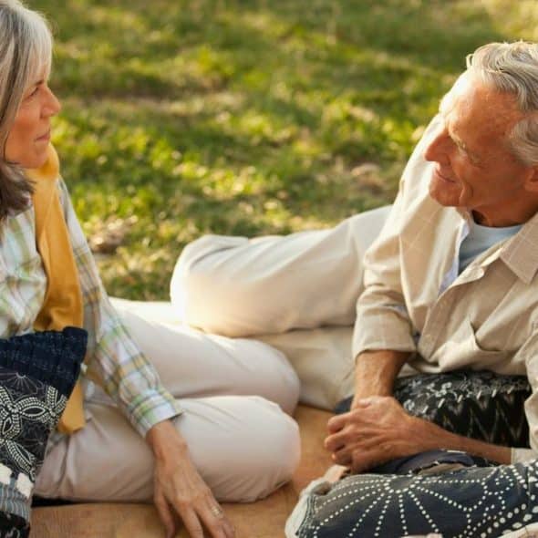 Elderly couple sitting on patterned pillows on a blanket outdoors in grass.