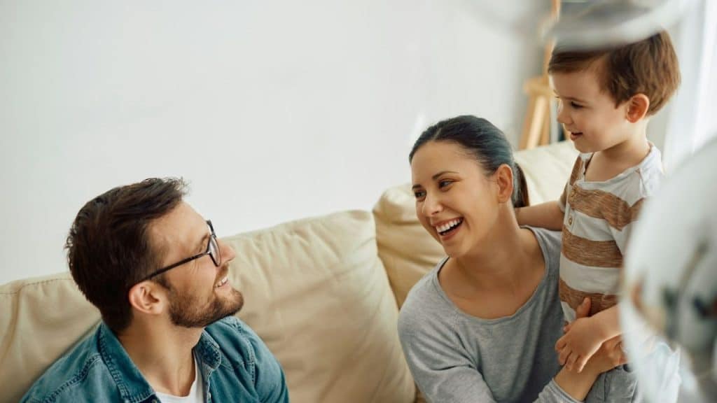 Smiling mother and son lean in on a couch towards a laughing father in glasses.