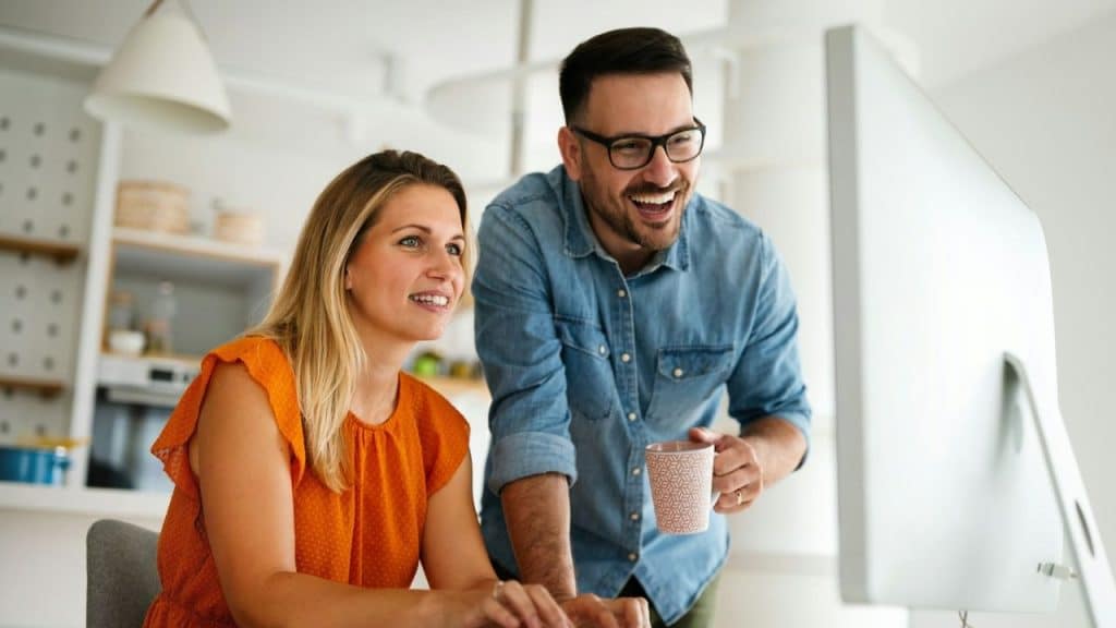 Smiling couple looks at computer monitor together, man stands holding a mug.